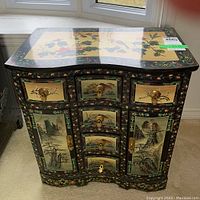 Front view of ornate black lacquered East Asian cabinet with curvy front, painted floral and landscape scenes, three drawers and two side doors with brass knobs.