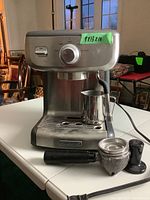 Front view of Calphalon espresso maker with portafilter, tamper, and milk frothing pitcher placed in front of the machine on table.