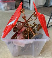 Plastic bin containing brass candle holders and candelabras loosely arranged, partially covered by plastic bin lid.