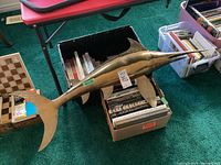 Full view of brass swordfish sculpture resting on cardboard boxes filled with books, showing entire length and fins.