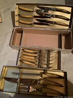 Multiple boxes laid open showing full set of knives and forks with light-colored handles, steel blades and tines, with original box showing some wear.