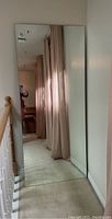 Full view of large, frameless wall mirror leaning against an upstairs wall near a staircase railing with beige curtains reflected in it.