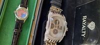 Two wrist watches side by side in original cases labeled Rayalty and Gastar respectively. Larger watch is silver-tone metal with rhinestone bezel and metal bracelet. Smaller watch has American flag dial and leather strap.