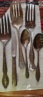 Photo of antique flatware set showing various forks, spoons, and a knife with heavy handles and ornate patterns with tarnish.