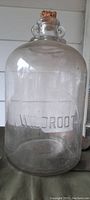 Large clear glass gallon jug embossed with 'WILDROOT', showing entire jug including cork stopper, handles on neck, and body shape.