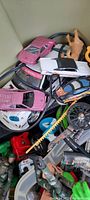 Photo showing pink and white die-cast model cars, some toy motorcycles, and mixed small figures in a plastic tub.