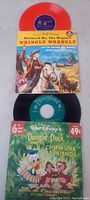 Two 7-inch vinyl records and their original sleeves. One black vinyl with Donald Duck and Chipmunks themed cover, one red vinyl with western cowboys themed cover.