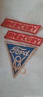 Three 1930s Ford dealer mechanics sewn patches on a plain background, one Ford V8 triangular patch and two Mercury rectangular patches.
