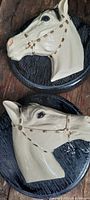 Two horse head wall decorations made of chalkware material with brown painted bridles on round wooden bases, placed on a rustic wooden surface.
