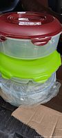 Plastic food storage containers primarily round, with colors including burgundy and green lids; containers are stacked and appear new with sealed lids.