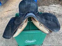 Top view of the black leather saddle showing seat and flaps with embossing and signs of wear, resting on a green plastic container outdoors.