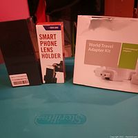 Photo showing the smartphone lens holder box, wireless earphones case, and the world travel adapter kit box on a blue surface.