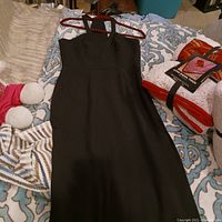 Full front view of the black sleeveless slim dress hanging on a red hanger, laid on a patterned bedspread with other household items around.