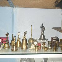 Shelved display of brass and metal bells with miniature decorative items including clocks, hourglass, candlestick, and small containers.