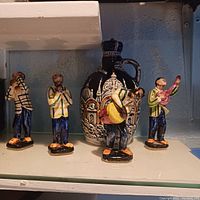 Four hand-painted ceramic musician figurines positioned in front of a decorative jar on a shelf, showing details of paint and posture.