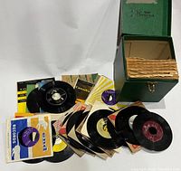 Photo showing 45 RPM vinyl records in original paper sleeves arranged beside a green vinyl record storage box with internal compartments.