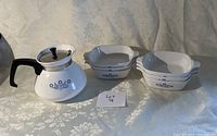 Vintage Corning Ware tea pot with blue cornflower design and six matching small dishes on a light floral tablecloth.