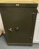 Front view of large green metal safe closed, showing combination dial and metal handle on door with visible hinges on right side.
