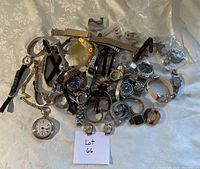 Multiple watches, watch bands, a pocket watch, and watch parts spread on a patterned fabric surface with a paper labeled 'Lot 66'.
