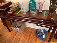 Full view of the wood console table with several decorative glass items on top and cardboard box and blue basket under the table.
