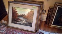 Overview of stacked framed paintings leaning against wall showing top painting with silver wood frame and forest river scene.