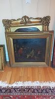 Two wood-framed art prints leaning against wall, shows one tavern scene print and empty carved frame behind