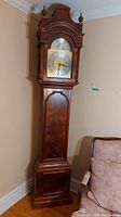 Full view of tall wooden grandfather clock with decorative top and lower panel door closed