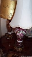 Close-up photo showing two decorative table lamps with porcelain bases featuring pink floral designs, gold accents, and white fabric lampshades with scalloped edges.