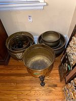 Six metal plant pots in corner, five matching brass-colored pots with claw-foot legs and one darker metal pot.