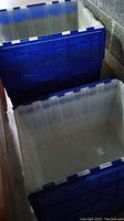 Two stacks of large plastic storage totes with blue bases and semi-transparent clear sides, some have lid flap latches visible, located next to a concrete block wall in a garage.