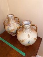 Photo showing two matching hand painted Austrian vases with floral motifs and gold accents on a wooden surface.
