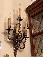 Pair of ornate bronze-colored metal wall sconces with five candle-like bulbs each mounted on a wall near a wood-framed window.