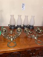Four battery operated lights with frosted glass shades and two brass wall hanging candle holders arranged on a wooden table.