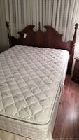 Photo of assembled bed showing dark wooden headboard with carved design and turned posts, mattress and box spring with tufted pattern, placed in bedroom setting with curtain backdrop.