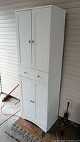 White tall cabinet made of press wood with two sets of doors and a central drawer. The cabinet is placed on a porch against a siding wall.