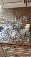 Overview photo showing multiple clear glass candle holders, a large clear glass vase, and two ivory battery operated candles on a kitchen counter.