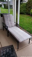 Adjustable metal frame chaise patio lounger with striped cushioned seat and backrest positioned on a porch with grass and a driveway in the background.
