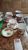 Seven assorted tea cups with matching saucers displayed on counter, showing various classic floral patterns including Castle China and Royal Albert Sea to Sea patterns.