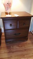 Front view of one of the wooden night tables showing its flat top, two small drawers with round knobs, and one larger drawer with ornate handle, placed on wood flooring with visible scratches on surface.