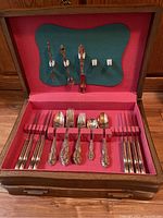 Flatware set displayed inside an open wooden box with red felt lining and green felt on the lid holding additional utensils.