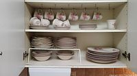 Shelves with white ceramic Gibson dinnerware including stacked dinner plates, side plates, bowls and hanging teacups with pink rose floral pattern.