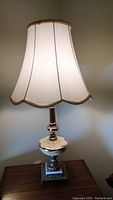 Full view of lamp with shade, showing tall white fabric shade with gold trim and ornate brass and stone base.