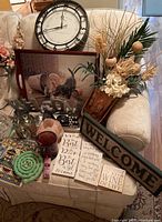 Photo showing the entire lot on a cream couch including a large wall clock, tray with dog image, welcome and inspirational signs, salt candle holder, and floral arrangement.