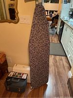 View of ironing board with paisley pattern cover standing upright next to vintage travel case and sewing kit on floor.