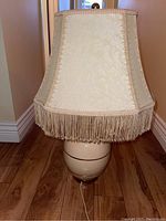 Full view of one vintage ceramic table lamp with cream fabric shade and decorative fringe trim on bottom edge, showing overall style and color.