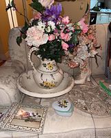 Photo showing entire grouping including ceramic washstand pitcher and bowl with floral pattern, shoe vase with dried flowers, crystal plate, floral trivets and heart-shaped trinket box, arranged on a cushioned armchair