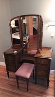 Front angle view of vintage vanity with three folding mirrors and upholstered stool in a room setting.