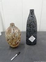 Two vintage vases side by side on a dark surface, showing one rounded cream and brown marbled vase and one tall black vase with floral design.