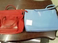 Two handbags on a brown table; a red leather Lancel handbag and a light blue Furla handbag labeled with number 54.