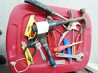 Top-down photo of a red storage bin showing a collection of various used hand tools in mixed conditions, including pliers, screwdrivers, hammers, wrenches, and cutters.